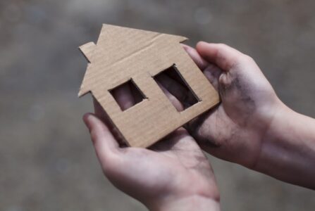 Et par hænder holder et lille lille hus skåret ud i pap.
