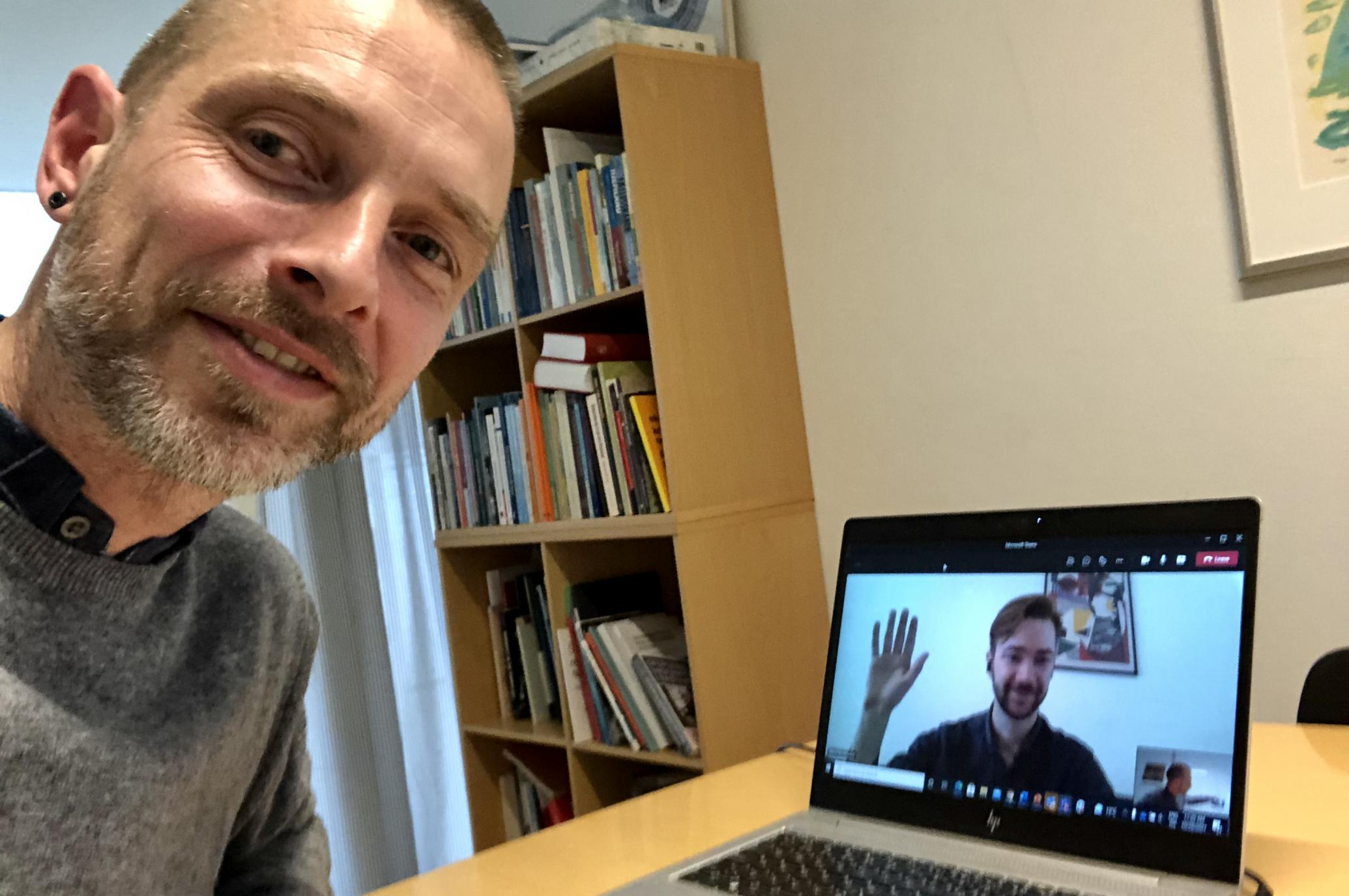 En mand sidder foran en laptop. På skærmen ses en anden mand, der vinker.