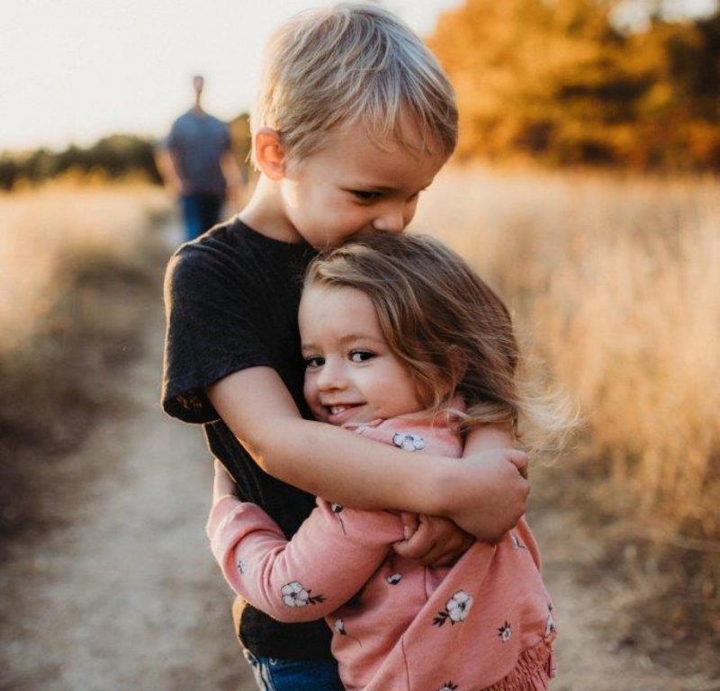 To mindre børn står og krammer hinanden på en sti. I baggrunden står en voksen.