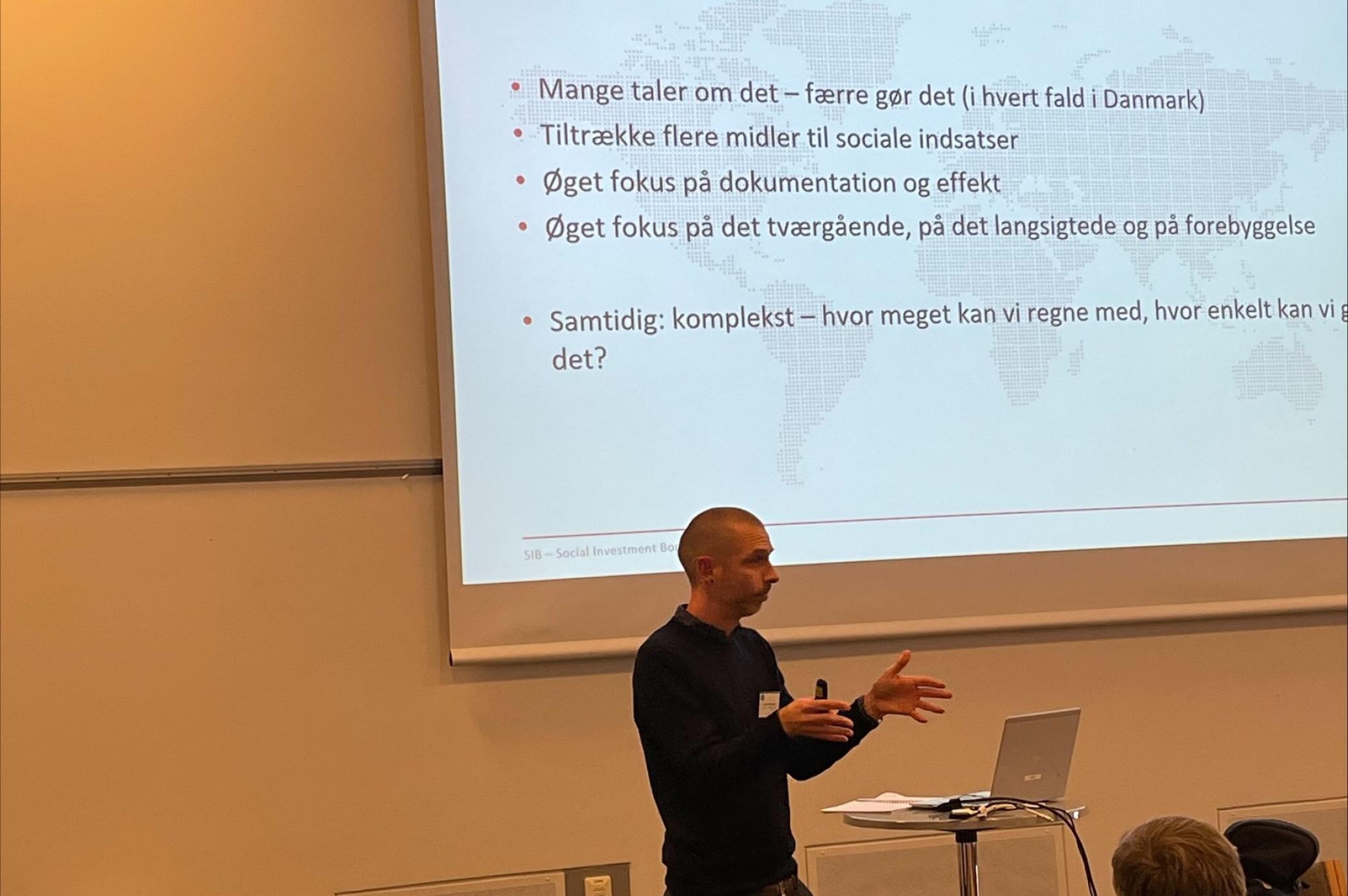 En person holder et oplæg - står ved en laptop og har et powerpoint slide på en skærm i baggrund.